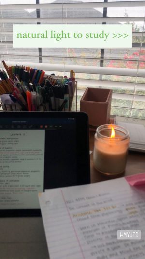 A desk top in front of a window. On the desk is a caddy of markers and pens, a tablet, sheets of note paper covered in notes, and a lit candle./