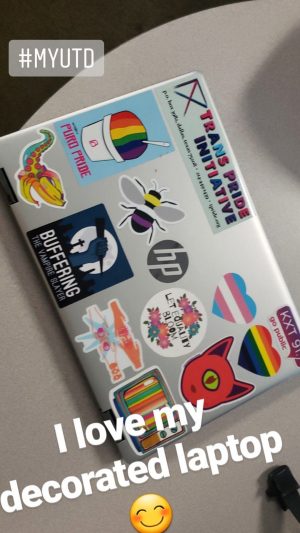 Emoji smiling with rosy cheeks. The cover of an HP laptop covered with stickers, inclusive ones that read, Trans Pride Initiative, Puro Pride, KXT Go Public, Let Equality Bloom, and Buffering: The Vampire Slayer. 
