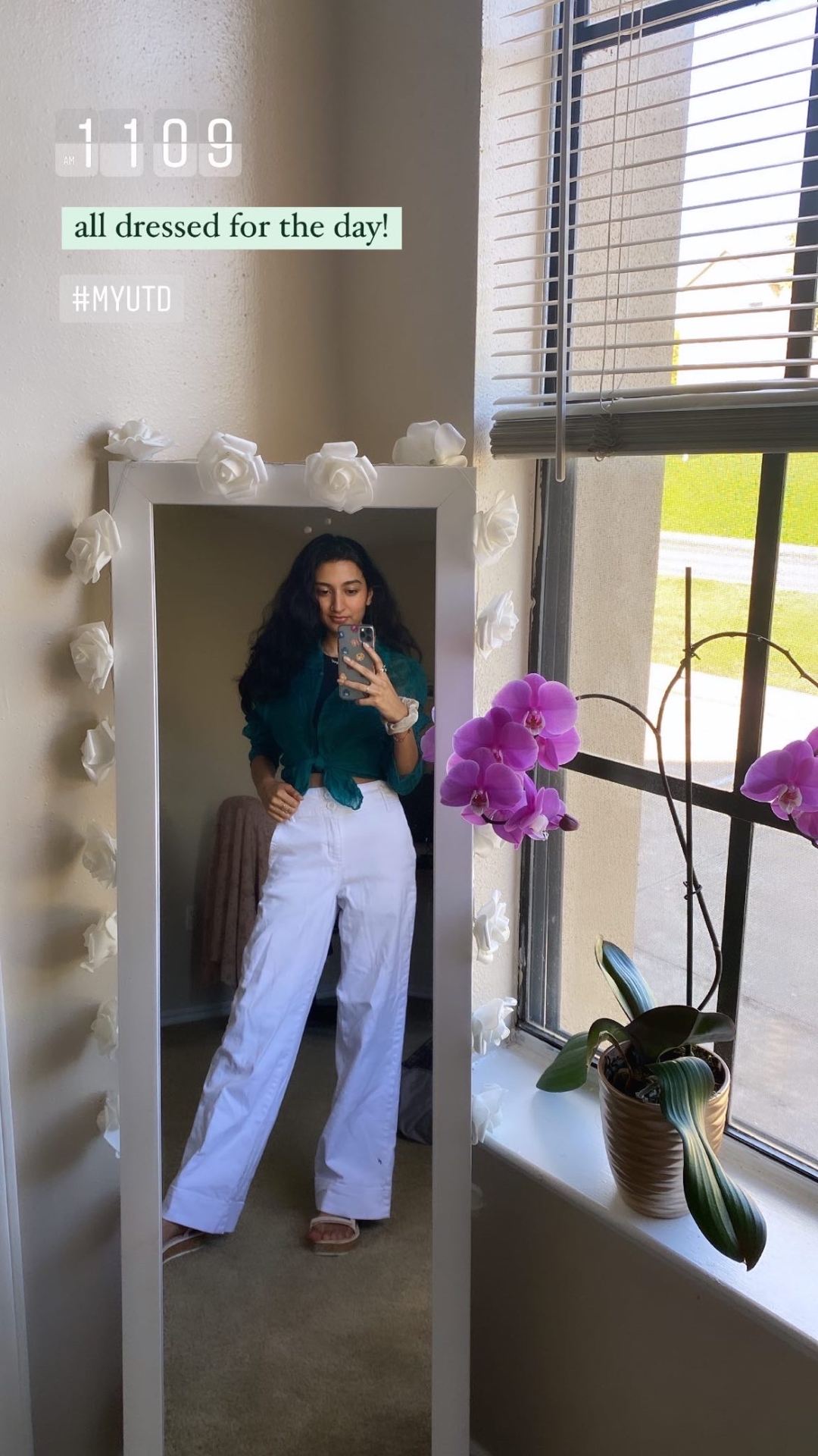 Twinkle takes a selfie, view in a mirror surrounded by white flowers. Twinkle wears a green top and white pants. There is a blooming pink orchid on a window sill. 