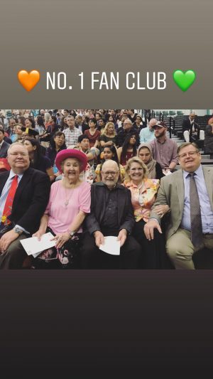A group of five people seated together in the graduation viewing room. They're on stadium seats with a crowd of other spectators behind them. Orange and green hearts surround the text, no. 1 fan club.