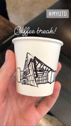 Rowan's hand holding a disposable espresso cup on which is a hand-drawn illustration of the ATEC building. 