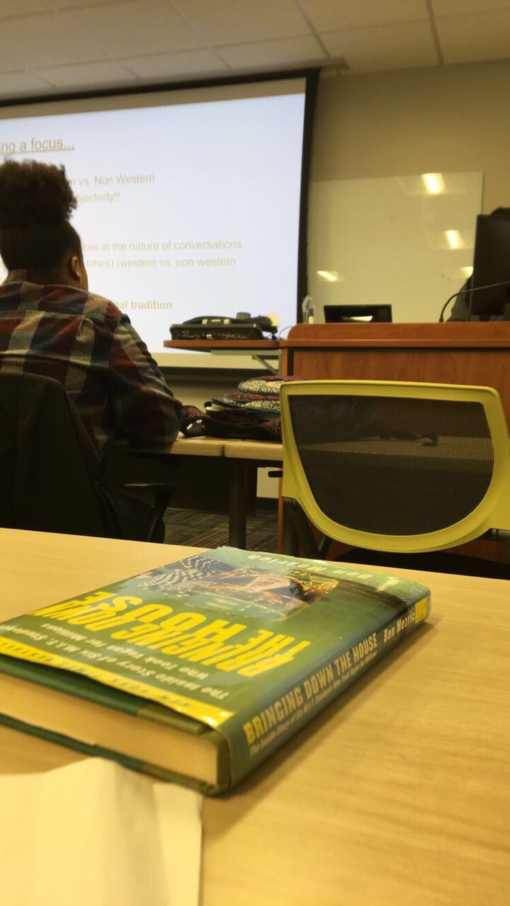 Ragya's point of view in class. Another student can be week, there's a book on a desk, and text is displayed on a presentation screen.